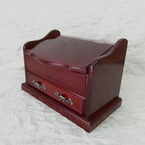 May include: A dark red wooden jewellery box. It has two drawers with decorative metal handles and a curved top. The box is designed in a classic style, ideal for storing jewellery or small keepsakes.