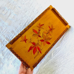 May include: A rectangular wooden tray with a light brown background and a floral design. The tray features a painted vine with red and orange leaves. The edges are decorated with a dark brown, patterned border. The tray is being held by a person.