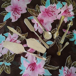May include: A collection of silver serving utensils, including cake servers, spoons, and a butter knife, are displayed on floral fabric. The utensils have decorative handles and are set against a backdrop of pink and blue flowers.
