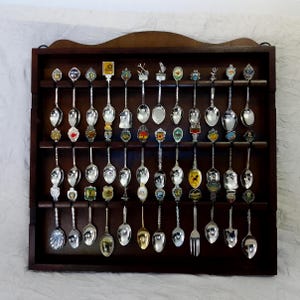 May include: A dark brown wooden wall-mounted display rack holding a collection of thirty-three silver-toned souvenir spoons. Each spoon features a unique decorative handle or emblem, arranged in three horizontal rows within the rectangular frame.