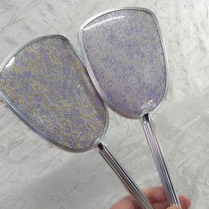 May include: Two vintage vanity sets, each featuring a brush and a mirror. The brush and mirror have a silver-toned frame and handle. The brush and mirror have a lavender and gold floral pattern.
