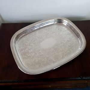 May include: A rectangular silver-toned serving tray featuring a polished finish, an intricate engraved floral pattern across the centre, a smooth oval-shaped blank space in the middle, and a decorative pierced gallery border with a ribbed edge.