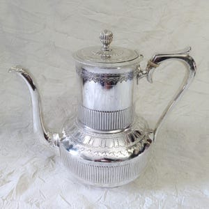 May include: A silver teapot with a decorative design and a lid with a knob. The teapot has a spout and a handle.