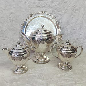 May include: A silver tea set with a tray. The set includes a teapot, a creamer, and a sugar bowl. Each piece has a decorative grape vine design and sits on a pedestal base. The tray has a scalloped edge.