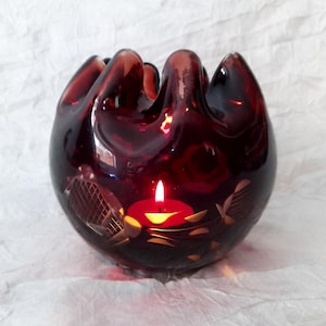 May include: A dark red glass candle holder with a lit candle inside. The candle holder has a distinctive shape with a ruffled top and etched floral patterns. The candle flame glows, creating a warm atmosphere.