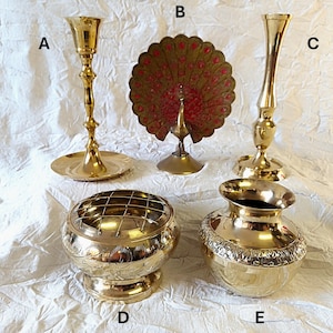 May include: A collection of brass ornaments for the home, featuring a tall candlestick holder, a peacock-shaped ornament, a slender vase, a floral frog, and a decorative vase with an ornate design. The items are displayed on a white surface.
