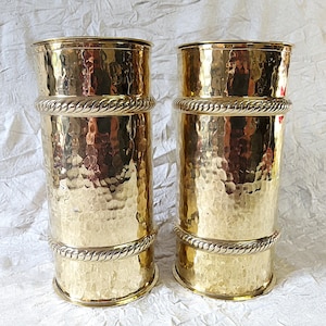 May include: Two matching gold-coloured metal vases with a hammered texture. Each vase has a cylindrical shape with a decorative rope-like band around the middle and base. The vases are displayed against a white background.