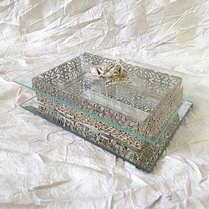 May include: A rectangular silver vanity tray with a glass top and a decorative rose. The tray has intricate filigree detailing and a mirrored base. The design creates a vintage aesthetic, suitable for a dressing table.
