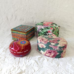 May include: Four decorative boxes: one square box with intricate orange, blue, and green geometric patterns; one hexagonal box with a pink and green floral print; one oval box with a pink and green floral print; and one round red box with painted floral designs and the text Playa del Carmen Mexico.