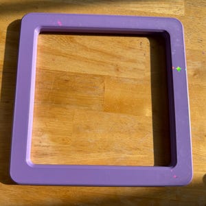 May include: A square, light purple frame with rounded corners and a smooth, flat surface. The frame features small, subtle, multi-colored star-shaped accents scattered along its edges, providing a simple, clean, and uniform border design.