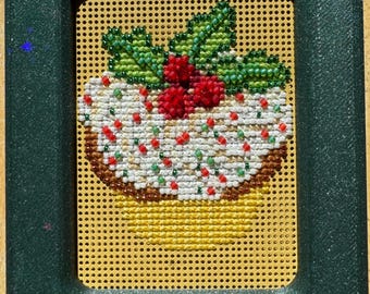 May include: A rectangular dark green glittery frame holds a perforated board featuring a beaded design of a holiday pudding. The pudding has white icing, red berries, and green holly leaves, with a yellow base. A metal needle rests at the top.