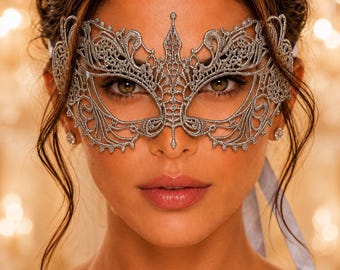 May include: A silver lace masquerade mask featuring an intricate filigree pattern with pointed edges and a central peak. The mask is secured with light gray ribbons tied at the sides, showcasing a detailed, openwork design that covers the eye area.