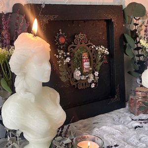 Puede incluir: Una vela de busto blanca con una llama encendida se encuentra junto a una exhibición enmarcada con un pequeño frasco. La exhibición está decorada con vegetación y pequeñas flores blancas. También se ve una pequeña vela en un portavelas de vidrio y un frasco de flores secas.