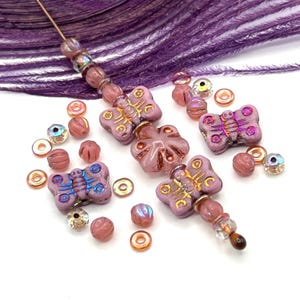 May include: A collection of pink and iridescent beads, including butterfly-shaped beads with gold accents, round beads, and small ring-shaped beads. A few beads are strung on a wire. The background includes purple feathers.