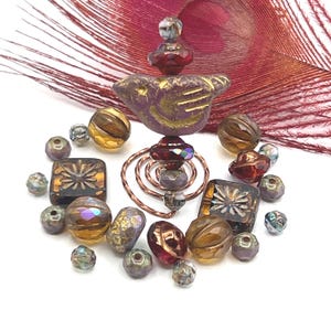 May include: A collection of artisan beads in various shapes and colors. The beads include faceted, square, and round designs in shades of purple, gold, and brown. A copper wire spiral and a decorative bird-shaped bead are also present, along with a maroon feather.