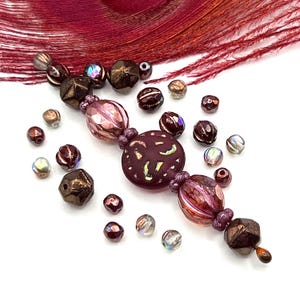 May include: An assortment of beads in various shapes and sizes, featuring shades of brown, purple, and iridescent finishes. The beads are scattered on a white surface, with a maroon feather in the background. The beads are suitable for jewelry making.