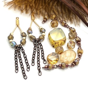 May include: An assortment of jewelry components, including two pairs of earrings with gold-colored wire and chain tassels, and a necklace with various beads in shades of gold, brown, and iridescent colors. The components are arranged on a white surface.