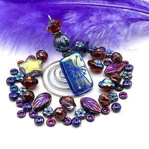 May include: A handcrafted bookmark with a blue and iridescent rectangular charm featuring a dandelion design. The bookmark is adorned with various beads in shades of blue, purple, and red, including star-shaped and flower-shaped beads. A purple feather is in the background.