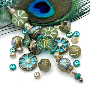 May include: An assortment of artisan beads in various shapes and colors. The beads include green, gold, and blue hues, with floral and leaf designs. A peacock feather is in the background.