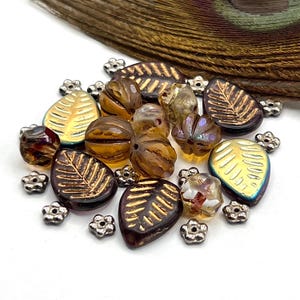 May include: A collection of glass beads in various shapes and colors. The beads include leaf-shaped, round, and faceted designs in shades of brown, gold, and iridescent hues. Small silver flower-shaped beads are scattered among them. A peacock feather is in the background.