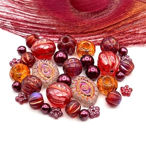 May include: A collection of glass beads in shades of red, orange, and purple. The beads vary in shape and size, including round, faceted, and floral designs. A maroon feather is in the background.