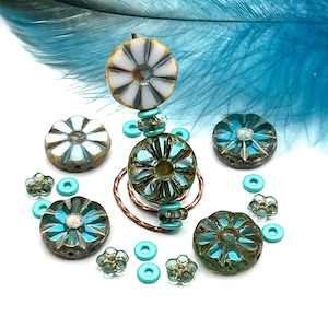May include: A collection of artisan beads in various shapes and sizes. The beads feature a mix of colors, including teal, white, and gold. Some beads have a floral design. A blue feather is in the background.
