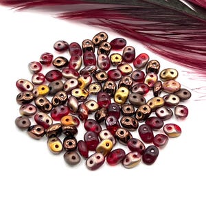 May include: A collection of small, oval-shaped beads in shades of red, gold, and bronze. The beads have two holes for stringing and are scattered on a white surface. A maroon feather is in the background.