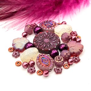 May include: A collection of various purple and iridescent glass beads, including round, floral, and snowflake designs. Accented with small copper-colored rings and a magenta feather in the background. Ideal for jewelry making.