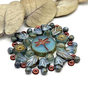 May include: A collection of artisan beads in various shapes and colors. The assortment includes blue, gold, and copper-toned beads, with a central round bead featuring a dragonfly design. The beads are arranged on a white surface.