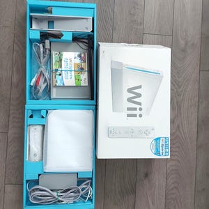 May include: A white Nintendo Wii video game console set in its original packaging. The box includes a white console, a Wii Remote controller, a Wii Sports game disc, a power adapter, and various connection cables, all organised in blue cardboard inserts.