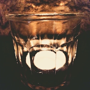 May include: A clear glass candle holder with a single white candle burning inside. The candle holder is sitting on a dark brown surface.