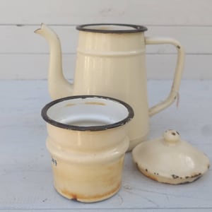 Può includere: Una caffettiera vintage in smalto crema con bordo nero e un coperchio e una tazza in smalto crema abbinati. La caffettiera ha un manico e un beccuccio. La tazza ha un bordo nero e un'iscrizione blu.