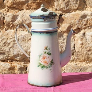 Könnte beinhalten: Vintage weiße Emaille-Kaffeekanne mit floralem Rosendesign. Die Kanne hat eine hellblaue Zierleiste und einen Deckel, einen gebogenen Ausguss und einen Griff. Die Kanne steht auf einer rosa Oberfläche.