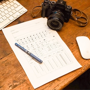 May include: A white sheet of paper with a grid of boxes and text. The paper is on a wooden table with a camera, a pen, and a computer mouse.