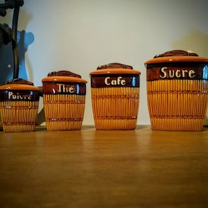 Puede incluir: Conjunto de cuatro botes de cerámica marrón con un patrón tejido. Los botes están etiquetados con "Poivre", "The", "Cafe" y "Sucre".