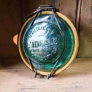 Op de afbeelding: Een vintage groene glazen flessenstop met een metalen draad sluiting. De dop heeft de tekst "Bouteilles L'Ideale by S.G.G.G. Paris" erop gedrukt.