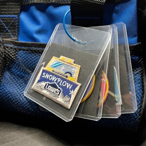 May include: Clear plastic holders containing embroidered patches, including a "SNOWPLOW LOWE'S" patch. The patches are displayed against a black background, with a blue cord attached to the holders. The background includes a blue and black bag.