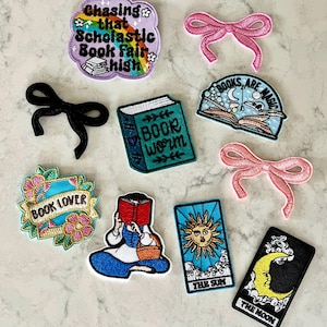 May include: Assortment of colorful iron-on patches featuring book-themed designs, including "Chasing that Scholastic Book Fair High", "Books are Magic", "Book Worm", "Book Lover", and tarot card-inspired patches with the Sun and Moon.