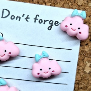 May include: Pink cloud shaped push pins with a blue bow and a smiley face. The push pins are on a white lined notepad with the text "Don't forget" written at the top.