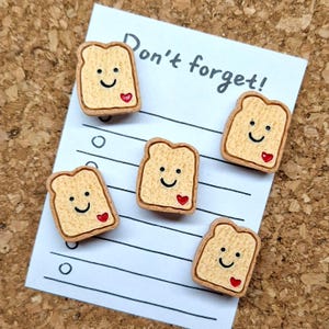 May include: Five smiling toast-shaped pins with a red heart and a happy face are arranged on a white notepad with the words "Don't forget!" written in black. The pins are tan with brown edges.
