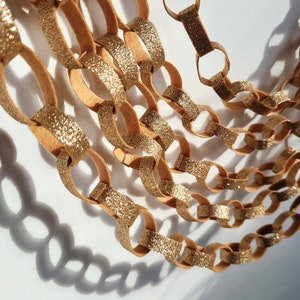 Könnte beinhalten: Nahaufnahme einer dekorativen Kette aus ineinandergreifenden Schlaufen. Die Kettenglieder wechseln sich zwischen einer beigefarbenen Farbe und einer goldglitzernden Oberfläche ab. Die Kette wirft einen Schatten auf eine weiße Oberfläche.