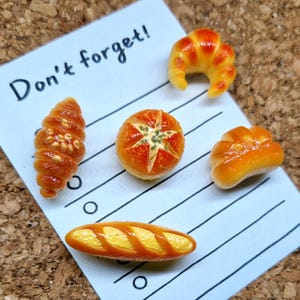 May include: An assortment of miniature baked goods, including croissants, a round bread, and a baguette, arranged on a lined paper with the text "Don't forget!" The baked goods are in shades of brown and yellow.