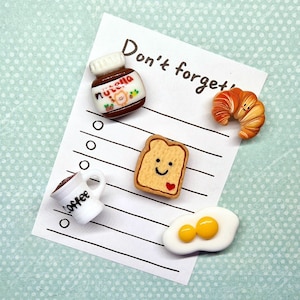 May include: A collection of miniature food items, including a Nutella jar, croissant, slice of toast with a heart, fried egg, and a coffee mug. The items are arranged on a list with the words "Don't forget" written at the top.