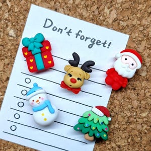 Puede incluir: Una lista de verificación con el texto "¡No te olvides!" escrito en ella. Hay cinco decoraciones navideñas en la lista de verificación: una caja de regalo roja y verde, un reno, un muñeco de nieve, un árbol de Navidad y Papá Noel.