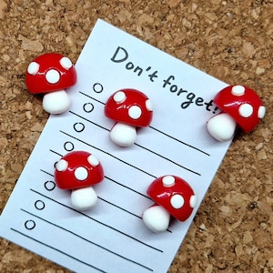 May include: Five red and white mushroom shaped push pins on a white lined notepad with the text "Don't forget!" written on it.