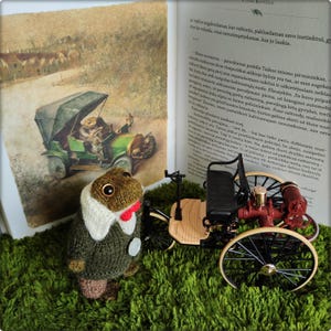 May include: A knitted frog doll wearing a green sweater, white collar, and red bow tie, standing next to a vintage toy car and an open book with an illustration of a frog driving a car.