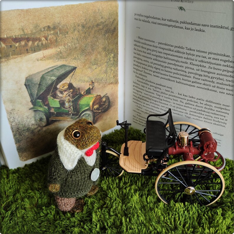 May include: A knitted frog doll wearing a green sweater, white collar, and red bow tie, standing next to a vintage toy car and an open book with an illustration of a frog driving a car.