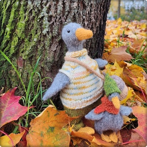 May include: A hand-knitted grey duck toy with an orange beak, wearing a cream-coloured jumper with thin yellow and blue stripes, standing next to a smaller grey duckling wearing a green hat and an orange scarf, both made of yarn.
