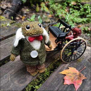 May include: A handcrafted, knitted frog figurine wearing a green tweed jacket, red bow tie, and white collar, standing next to a miniature antique car. The frog has large, expressive eyes and a button closure on its jacket.