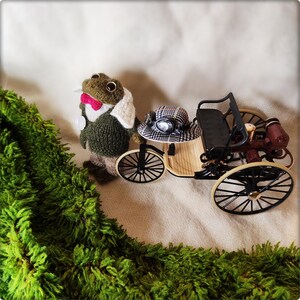 May include: A whimsical scene featuring a knitted frog wearing a jacket, bow tie, and a white collar, standing next to a miniature vintage car. The car has a black seat, wooden body, and black wheels. A green, textured border frames the scene.
